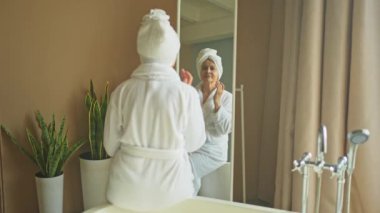 Yetişkin, yaşlı bir kadın aynaya bakıyor ve günlük güzellik kremi sürüyor. Cildinde beyaz maske kremi olan yaşlı bir kadın cilt bakımı görüyor.