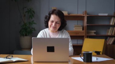 Evde dizüstü bilgisayar kullanan kendine güvenen Avrupalı orta yaşlı bir kadın. Yaşlı, şık 60 'lı yaşlarda bir kadın masada oturmuş bilgisayar ekranına bakıyor daktilo yazarak sohbet ediyor e-posta yazıyor.