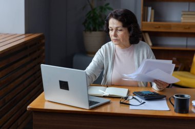 Orta yaşlı yaşlı bir kadın dizüstü bilgisayarı ve kağıt dökümanlarıyla oturuyor, evde kağıt faturalarını okuyan yaşlı bir kadın banka yöneticiliği finanse ediyor kredi emekliliği ödemelerini planlayan vergileri hesaplıyor.