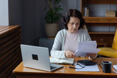 Orta yaşlı yaşlı bir kadın dizüstü bilgisayarı ve kağıt dökümanlarıyla oturuyor, evde kağıt faturalarını okuyan yaşlı bir kadın banka yöneticiliği finanse ediyor kredi emekliliği ödemelerini planlayan vergileri hesaplıyor.