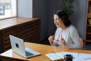 Orta yaşlı mutlu bir kadın dizüstü bilgisayarla oturup arkadaşlarıyla görüntülü konuşma yapıyor. Yaşlı ve olgun büyükanneme gülüp yetişkin çocuklarla internette konuşarak eğleniyorum.