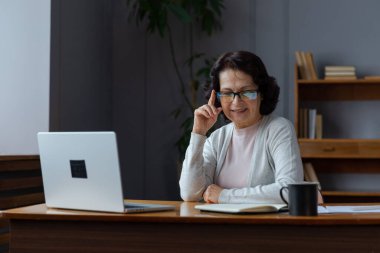 Evde dizüstü bilgisayar kullanan kendine güvenen Avrupalı orta yaşlı bir kadın. Yaşlı, şık 60 'lı yaşlarda bir kadın masada oturmuş bilgisayar ekranına bakıyor daktilo yazarak sohbet ediyor e-posta yazıyor.