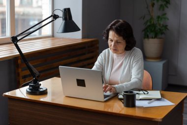 Evde dizüstü bilgisayar kullanan kendine güvenen Avrupalı orta yaşlı bir kadın. Yaşlı, şık 60 'lı yaşlarda bir kadın masada oturmuş bilgisayar ekranına bakıyor daktilo yazarak sohbet ediyor e-posta yazıyor.