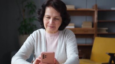 Avrupalı orta yaşlı bir kadın elinde akıllı telefon ekranı dokunmatik parşömen sayfası tutuyor. Evde internetteki sosyal medya uygulamalarını cep telefonuyla kullanan yaşlı ve şık bir kadın. İnternetten alışveriş