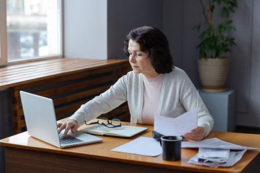 Middle aged senior woman sit with laptop and paper document. Pensive older mature lady reading paper bill pay online at home managing bank finances calculating taxes planning loan debt pension payment