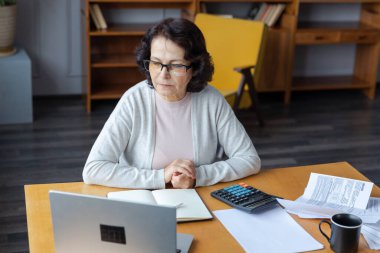 Middle aged senior woman sit with laptop and paper document. Pensive older mature lady reading paper bill pay online at home managing bank finances calculating taxes planning loan debt pension payment