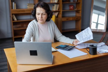 Middle aged senior woman sit with laptop and paper document. Pensive older mature lady reading paper bill pay online at home managing bank finances calculating taxes planning loan debt pension payment