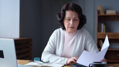 Middle aged senior woman sit with laptop and paper document. Pensive older mature lady reading paper bill pay online at home managing bank finances calculating taxes planning loan debt pension payment