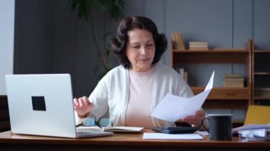 Middle aged senior woman sit with laptop and paper document. Pensive older mature lady reading paper bill pay online at home managing bank finances calculating taxes planning loan debt pension payment