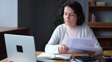 Orta yaşlı yaşlı bir kadın dizüstü bilgisayarı ve kağıt belgeleriyle oturuyor. Düşünceli, olgun, kağıt faturası okuyan, ev idaresi, banka hesapları, kredi emekliliği ödemesi planlayan vergileri finanse eden yaşlı bir kadın.