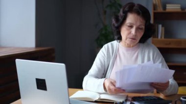 Middle aged senior woman sit with laptop and paper document. Pensive older mature lady reading paper bill pay online at home managing bank finances calculating taxes planning loan debt pension payment