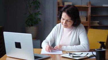 Çevrimiçi eğitim kursları webinar konsepti. Orta yaşlı yaşlı bir kadın dizüstü bilgisayar kullanarak not tutuyor. Odaklanmış yaşlı kadın çevrimiçi çalışmaktan zevk alıyor.