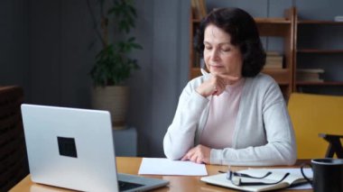 Evde dizüstü bilgisayar kullanarak not yazan orta yaşlı bir kadın. Yetişkin, yaşlı bir iş kadınının internet üzerinden kayıt yapması. Olgun patron profesyonel işçi.