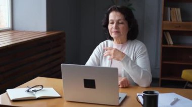 Evde dizüstü bilgisayar kullanan kendine güvenen Avrupalı orta yaşlı bir kadın. Yaşlı, şık 60 'lı yaşlarda bir kadın masada oturmuş bilgisayar ekranına bakıyor daktilo yazarak sohbet ediyor e-posta yazıyor.