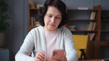 Avrupalı orta yaşlı bir kadın elinde akıllı telefon ekranı dokunmatik parşömen sayfası tutuyor. Evde internetteki sosyal medya uygulamalarını cep telefonuyla kullanan yaşlı ve şık bir kadın. İnternetten alışveriş