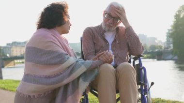 Tekerlekli sandalyedeki yaşlı adam parkta yaşlı bir kadınla yürüyor. Dışarıda özürlü insanlar için tekerlekli sandalyedeki felçli adamla ilgilenen yaşlı aile kadını. Rehabilitasyon