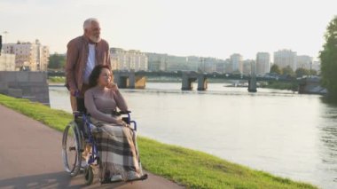 Tekerlekli sandalyede yaşlı bir adamla parkta yürüyen yaşlı bir kadın. Yaşlı aile erkeği koca, dışarıda çalışanlar için sandalyeye oturan kadını kucaklamayı destekliyor. Rehabilitasyon
