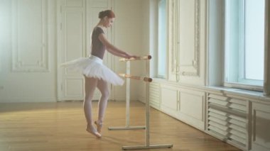 Tutu etekli genç balerin dans sınıfında bale barının yanında esneme hareketleri yapıyor. Dans stüdyosundaki genç klasik dansçı kadın. Bale sınıfı eğitimi. Bale okulu konsepti