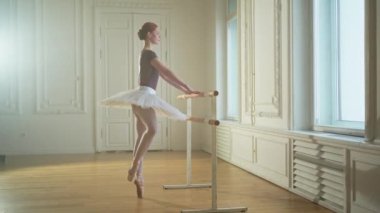 Tutu etekli genç balerin dans sınıfında bale barının yanında esneme hareketleri yapıyor. Dans stüdyosundaki genç klasik dansçı kadın. Bale sınıfı eğitimi. Bale okulu konsepti
