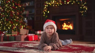 Mutlu noeller. Küçük çocuk klasik karanlık iç mekanda Noel ağacının yanında gülümsüyor. Genç mutlu çocuk Noel Baba şapkasını şömineli Noel ağacı hediye kutularıyla oturma odasında giyiyor. Evde Noel arifesi