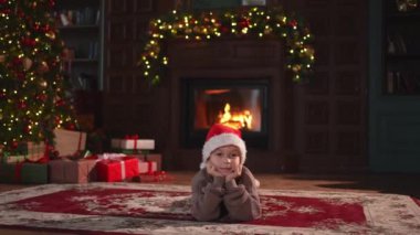 Mutlu noeller. Küçük çocuk klasik karanlık iç mekanda Noel ağacının yanında gülümsüyor. Genç mutlu çocuk Noel Baba şapkasını şömineli Noel ağacı hediye kutularıyla oturma odasında giyiyor. Evde Noel arifesi