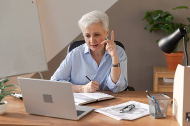 İş yerinde dizüstü bilgisayar kullanarak not yazan orta yaşlı bir kadın. Yetişkin, yaşlı bir iş kadınının ofiste çalışıp, online işler yaparak yazılı plaklar çıkarmasına odaklanmış. Olgun patron profesyonel işçi.