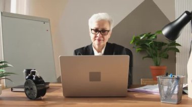 İş yerinde dizüstü bilgisayar kullanan kendine güvenen Avrupalı orta yaşlı bir kadın. Modaya uygun, olgun 60 'ların beyaz saçlı iş kadını ofis masasında oturuyor. Patron öğretmen profesyonel işçi.
