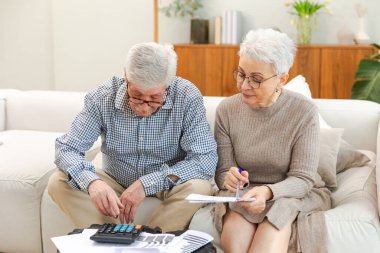Orta yaşlı yaşlı çift dizüstü bilgisayarları ve kağıt belgeleriyle oturuyor. Yaşlı, olgun bir kadın kağıt faturası okuyor. Ev yöneticiliği yapıyor. Vergileri finanse ediyor.