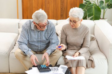 Orta yaşlı yaşlı çift dizüstü bilgisayarları ve kağıt belgeleriyle oturuyor. Yaşlı, olgun bir kadın kağıt faturası okuyor. Ev yöneticiliği yapıyor. Vergileri finanse ediyor.