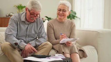 Orta yaşlı yaşlı çift kağıt döküman ile oturup mali evrak işleriyle uğraşıyor. Yaşlı olgun bir kadın kağıt faturası okuyor. Banka yönetiyor. Vergileri hesaplıyor.