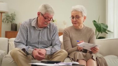 Orta yaşlı yaşlı çift kağıt döküman ile oturup mali evrak işleriyle uğraşıyor. Yaşlı olgun bir kadın kağıt faturası okuyor. Banka yönetiyor. Vergileri hesaplıyor.