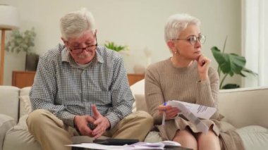 Orta yaşlı yaşlı çift kağıt döküman ile oturup mali evrak işleriyle uğraşıyor. Yaşlı olgun bir kadın kağıt faturası okuyor. Banka yönetiyor. Vergileri hesaplıyor.