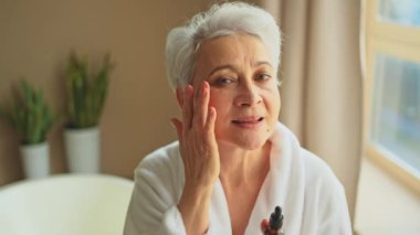 Cilde doğal damla kozmetik yağı süren bir kadın. Yetişkin yaşlı bir kadın günlük güzellik rutini yapan pipetli anti-yaş serumu uyguluyor. Yaşlı kadın yüz bakımı yaptırıyor.
