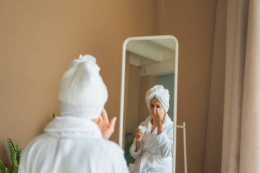 Yetişkin, yaşlı bir kadın aynaya bakıyor ve günlük güzellik kremi sürüyor. Cildinde beyaz maske kremi olan yaşlı bir kadın cilt bakımı görüyor.