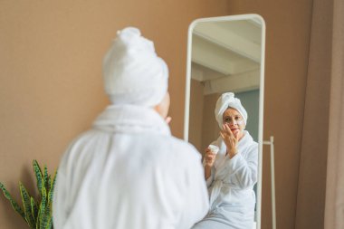 Yetişkin, yaşlı bir kadın aynaya bakıyor ve günlük güzellik kremi sürüyor. Cildinde beyaz maske kremi olan yaşlı bir kadın cilt bakımı görüyor.