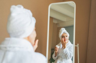 Yetişkin, yaşlı bir kadın aynaya bakıyor ve günlük güzellik kremi sürüyor. Cildinde beyaz maske kremi olan yaşlı bir kadın cilt bakımı görüyor.