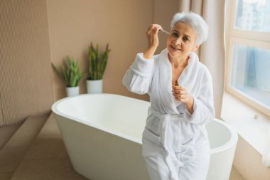 Cilde doğal damla kozmetik yağı süren bir kadın. Yetişkin yaşlı bir kadın günlük güzellik rutini yapan pipetli anti-yaş serumu uyguluyor. Yaşlı kadın yüz bakımı yaptırıyor.