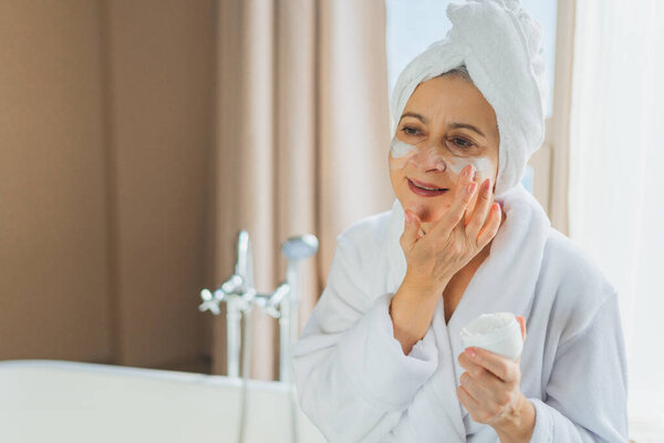 Mature adult old woman applying nourishing anti age face cream doing daily beauty routine. Senior woman with white mask or creme on skin gets facial treatment Skin care aging beauty skincare cosmetics