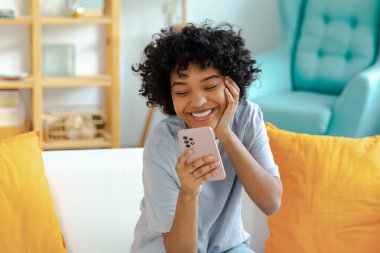 Cep telefonuyla gülen heyecanlı, mutlu, genç siyahi, Afro-Amerikalı kadın. Cep telefonu mesajı almaktan zevk alıyor. Neşeli kız yüksek sesle gülüyor. Kanepede oturup komik video haberleri izliyor.