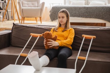 Kanepede oturan kırık bacaklı genç kız. Üzgün küçük çocuk, kırık bacağını iyileştirmek için oyuncak ayısına sarılıyor. Alçı ve koltuk değneği kullanan bir kız. Kaza sonucu oluşan kırık, ayak bandajı.