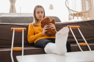 Kanepede oturan kırık bacaklı genç kız. Üzgün küçük çocuk, kırık bacağını iyileştirmek için oyuncak ayısına sarılıyor. Alçı ve koltuk değneği kullanan bir kız. Kaza sonucu oluşan kırık, ayak bandajı.