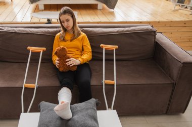 Kanepede oturan kırık bacaklı genç kız. Üzgün küçük çocuk, kırık bacağını iyileştirmek için oyuncak ayısına sarılıyor. Alçı ve koltuk değneği kullanan bir kız. Kaza sonucu oluşan kırık, ayak bandajı.