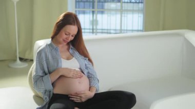 Hamilelik anneliği insanların gelecek beklentisi. Hamile bir kadın koca göbeğine dokunuyor. Evdeki koltukta oturuyor. Karnına sarılan ve hamileliğin tadını çıkaran bir kız. Annelik şefkati ebeveynlik yeni yaşam konsepti