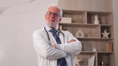 Gözlüklü yaşlı doktor hastane ya da doktor muayenehanesinde kameraya gülümsüyor. Pratisyen hekim, klinikte profesyonel sağlık uzmanı. Sağlık Hizmetleri