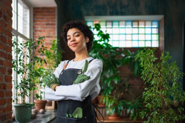 Kendi işimi yürütüyorum. Botanik mağazasında yeşil bitkilerle önlük giyen Afrikalı bir çiçekçi kadın. Çiçekçide çalışan mutlu küçük işletme sahibi etrafı bitkilerle çevrili gülümsüyor.