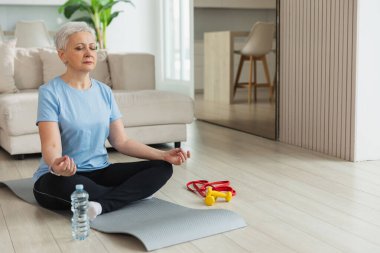 Yoga farkındalığı meditasyonu. Yetişkin bir kadın evde yoga yapıyor. Orta yaşlı kadın nilüfer çiçeğinde oturmuş yoga minderinde meditasyon yaparken poz veriyor. Yaşlı orta yaşlı kadın nefes egzersizi yapıyor.