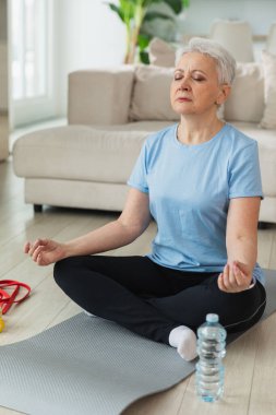 Yoga farkındalığı meditasyonu. Yetişkin bir kadın evde yoga yapıyor. Orta yaşlı kadın nilüfer çiçeğinde oturmuş yoga minderinde meditasyon yaparken poz veriyor. Yaşlı orta yaşlı kadın nefes egzersizi yapıyor.