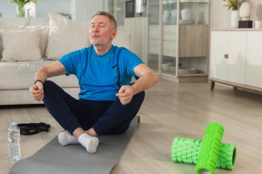 Yoga farkındalığı meditasyonu. Yetişkin bir adam evde yoga yapıyor. Orta yaşlı büyükbaba nilüfer çiçeğinde oturmuş yoga minderinde meditasyon yaparken poz veriyor. Yaşlı adam nefes alma pratiği yapıyor.