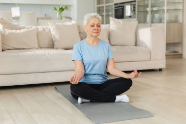 Yoga farkındalığı meditasyonu. Yetişkin bir kadın evde yoga yapıyor. Orta yaşlı kadın nilüfer çiçeğinde oturmuş yoga minderinde meditasyon yaparken poz veriyor. Yaşlı orta yaşlı kadın nefes egzersizi yapıyor.