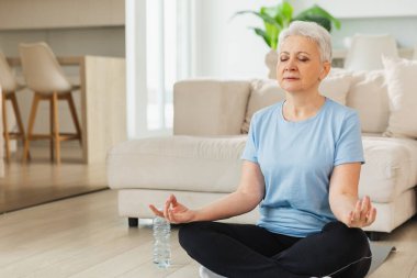 Yoga farkındalığı meditasyonu. Yetişkin bir kadın evde yoga yapıyor. Orta yaşlı kadın nilüfer çiçeğinde oturmuş yoga minderinde meditasyon yaparken poz veriyor. Yaşlı orta yaşlı kadın nefes egzersizi yapıyor.